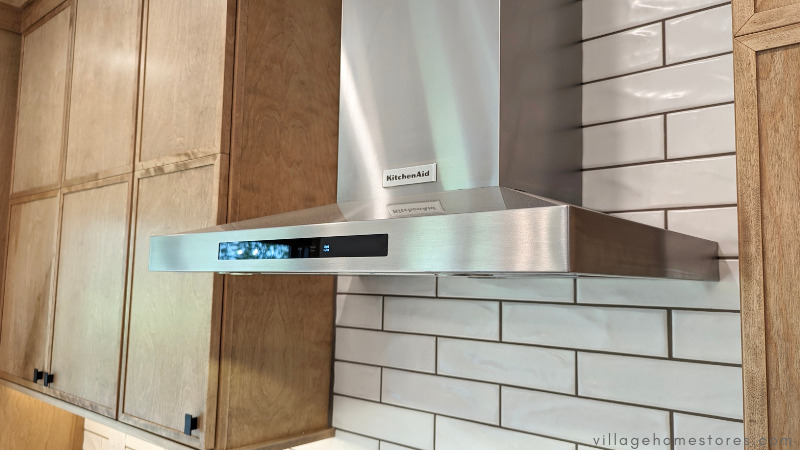 close up of stainless steel kitchenaid chimney style range hood against long white wall tile