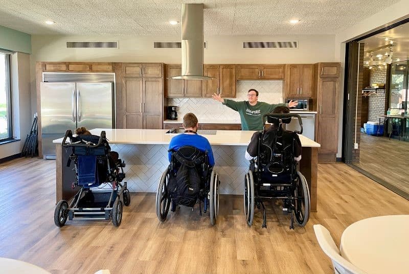 three wheelchair users rolled up to the kitchen island with young man holding hands open showing the kitchen