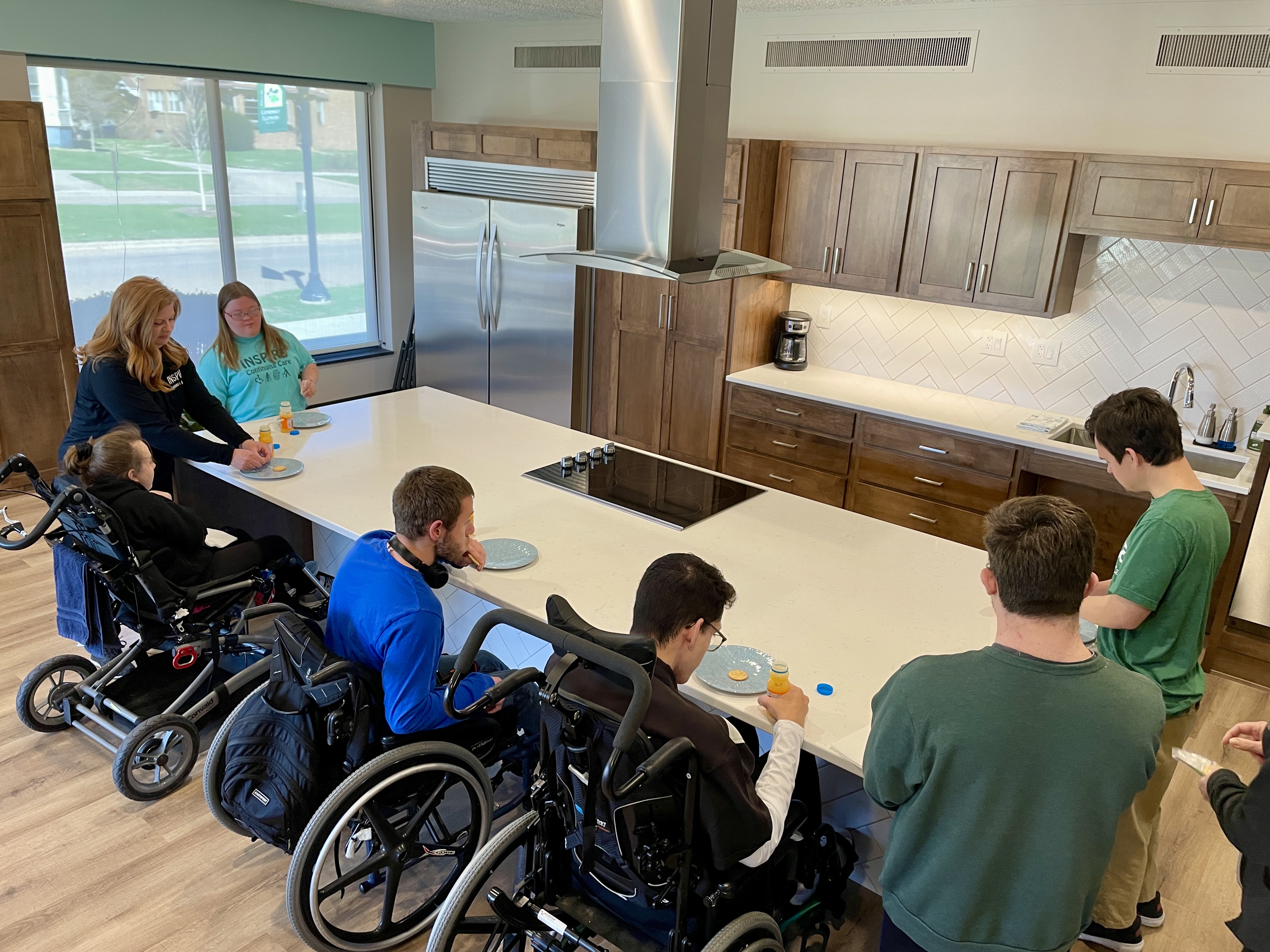 adults and young adults standing and in wheelchairs surround a large kitchen island with cooktop and hood above
