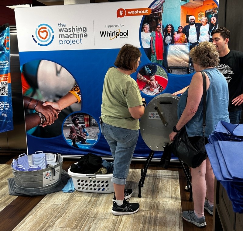 image of women at divya manual washing machine getting a demonstration with backdrop and laundry baskets on floor