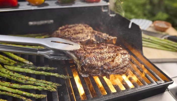 A pair of tongs flips a steak on a grill next to some asparagus.