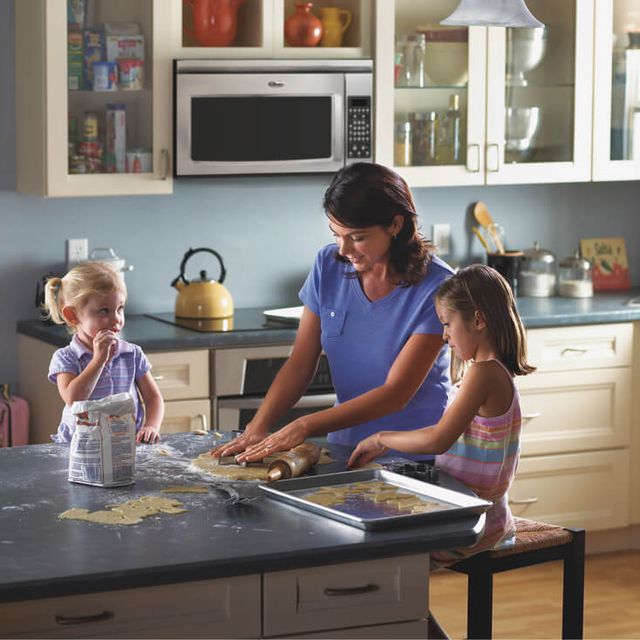 front view of a woman and two children in a traditional kitchen. The woman is demonstrating how to cut the cookie dough on the counter in front of her, as one of the girls watches and the other eats the dough!