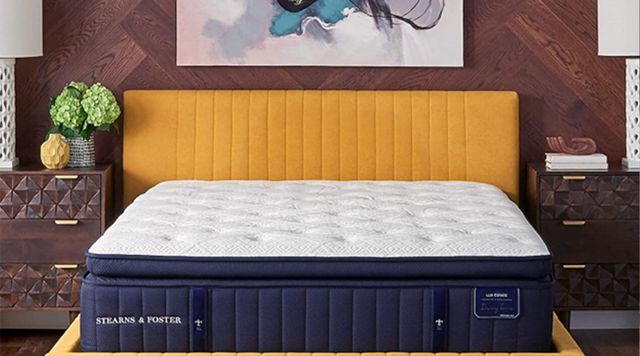 front view of a stearns and foster pillow top mattress in a modern bedroom with a yellow headboard, wood panel walls, and white and blue artwork above
