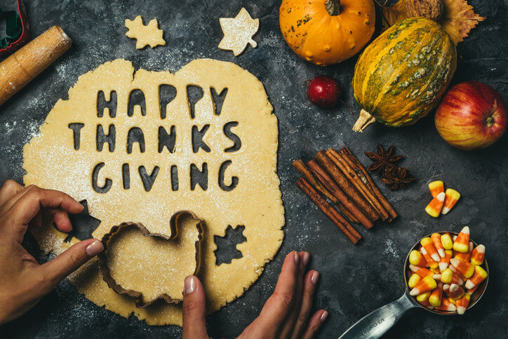 Image of pie dough being rolled out on a dark counter with pumpkins and leaf-shaped dough cutouts around the dough. The dough is flattened and the words "Happy Thanksgiving" are cut out of the dough