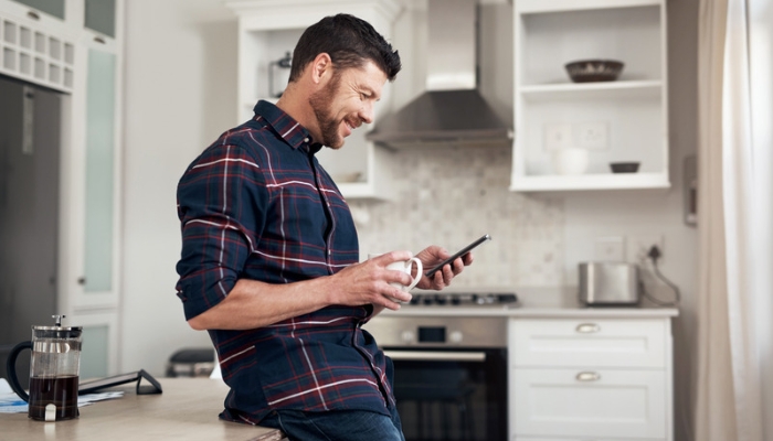 A man drinks coffee in his kitchen while using his cell phone