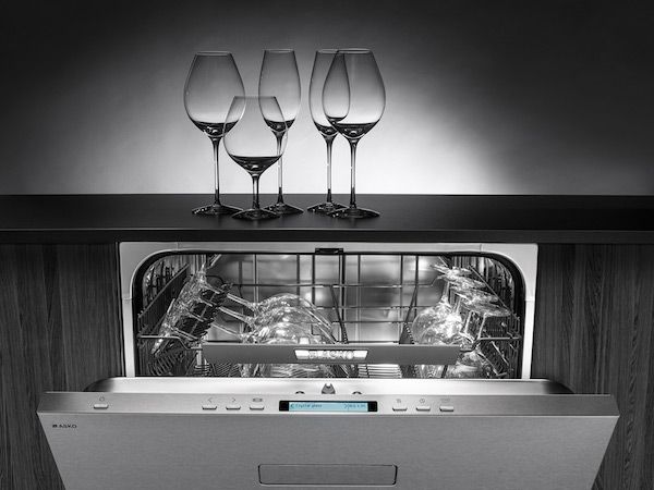 front view of an asko dishwasher built into black cabinetry, slightly opened to show it is full of wine glasses with various glassware on the counter above