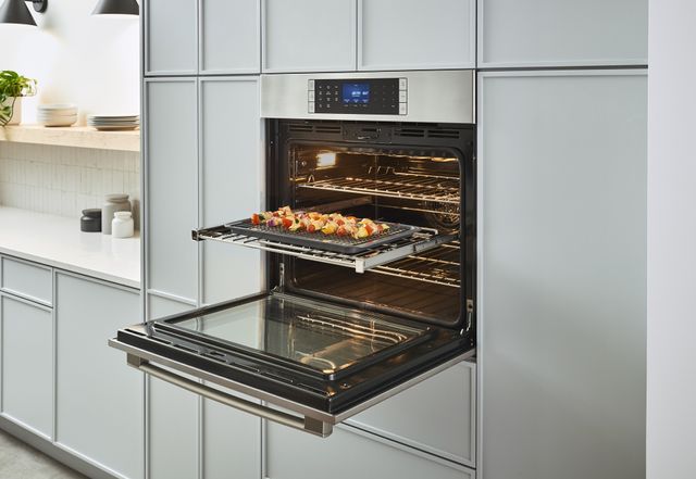 front view of an opened wall oven with a dish pulled out on the rack, and the oven is built into a wall of white cabinetry