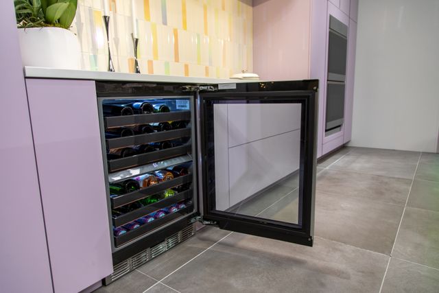 View of an opened under counter wine refrigerator stocked with wine in a modern kitchen with light pink cabinetry and white walls.
