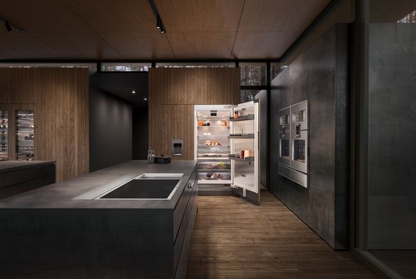 Front view of a contemporary kitchen with a Gaggenau cooktop in an island, an opened Gaggenau refrigerator, and wall ovens.