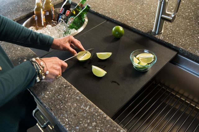 top view of two arms using a knife to cut limes on a cutting board in a workstation sink, with a pail of drinks in the sink to the left