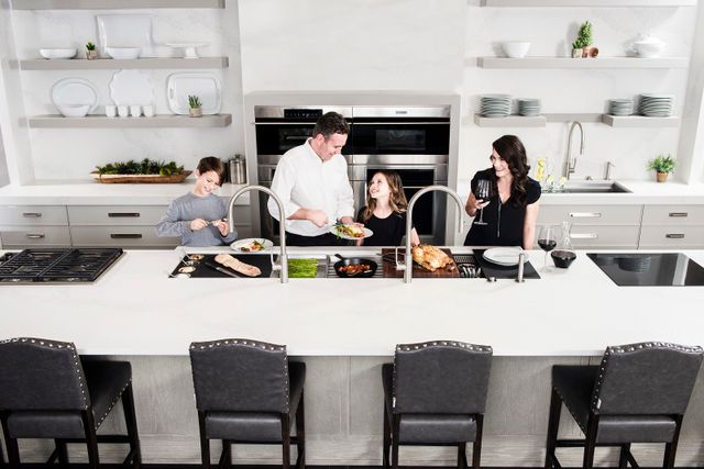 front aerial view of an all white kitchen with and island with a workstation sink and two parents and children preparing dinner around it