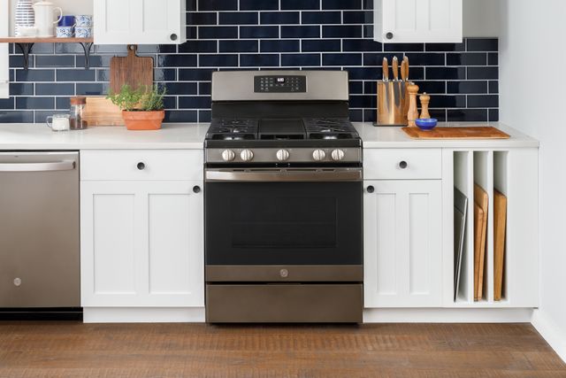 front view of a modern kitchen with white cabinets and countertops, blue subway tile backsplash, and stainless steel appliances including a range and dishwasher