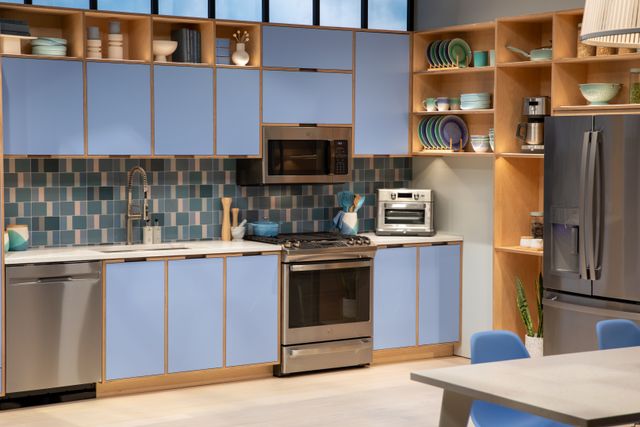 front view of a modern kitchen with blue and wooden cabinetry, a blue backsplash, and stainless steel appliances including a range, dishwasher, over the range microwave, and refrigerator 
