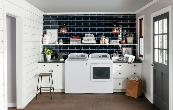 White GE top load washer and dryer in a laundry room with a blue brick wall