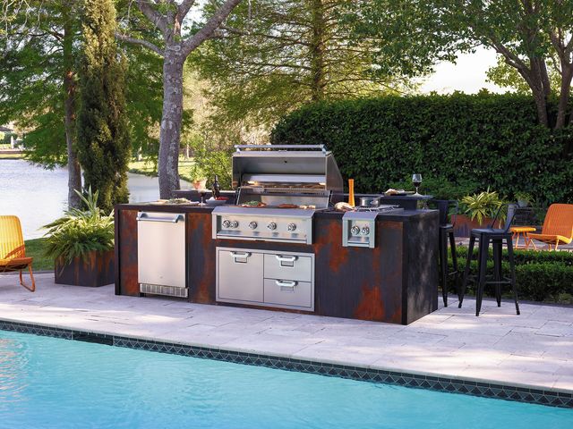 front view of a modern outdoor kitchen with an open natural gas grill with various meats cooking on it, and other outdoor kitchen appliances around