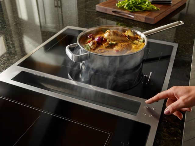 Front view of a JennAir downdraft cooktop with food cooking on top and steam being pulled into the vent