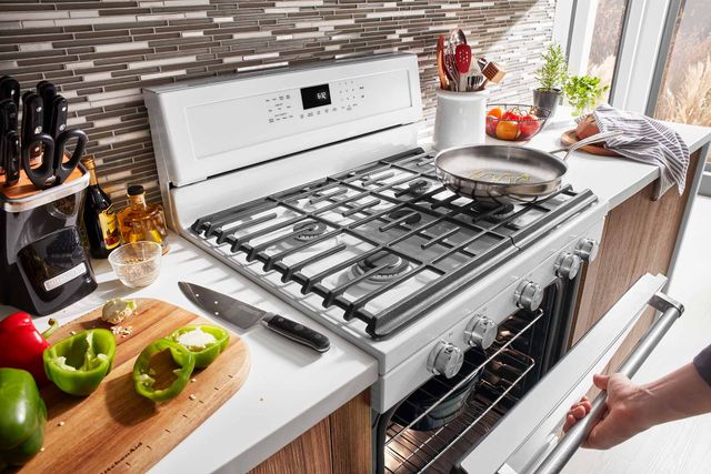 Close view of a KitchenAid 36" gas range with a stainless steel pan on top of the front right burner and a hand reaching to open the oven