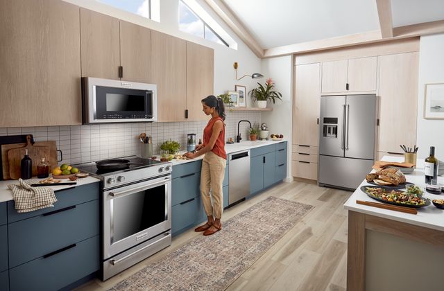 View of a modern kitchen with white walls, light wooden cabinetry, and stainless steel appliances. A woman prepares food in the kitchen next to the range.