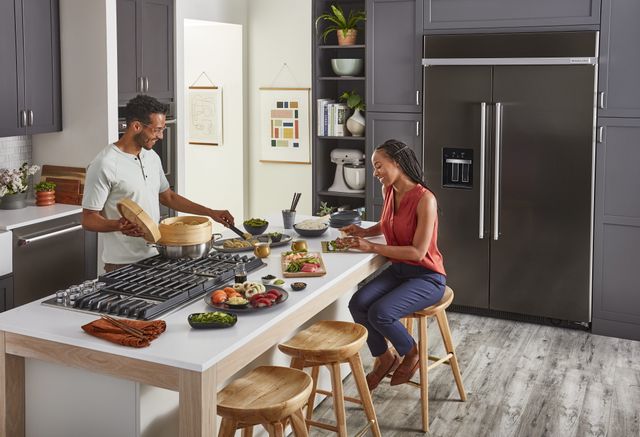 Front view of a modern kitchen with a man making dinner on a KitchenAid rangetop and a woman chopping veggies on a kitchen island
