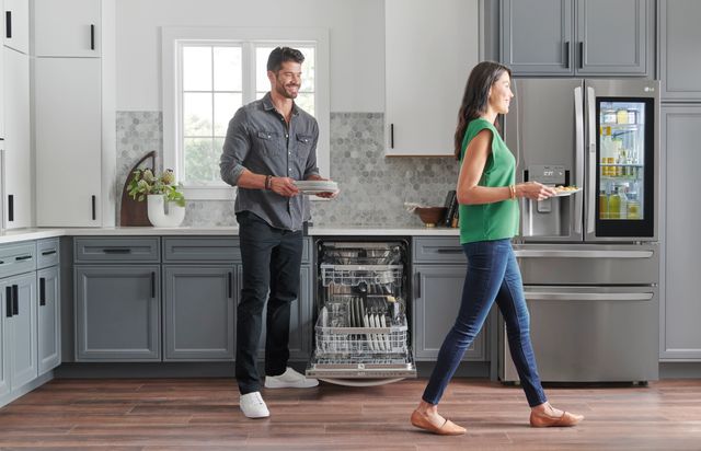 Front view of a couple smiling and unloading their LG dishwasher in a modern kitchen with an LG refrigerator as well.