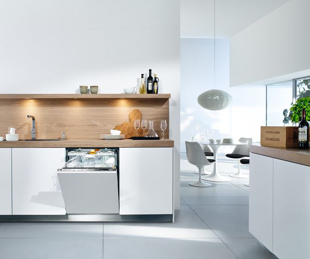 front view of a high end minimalist kitchen with white walls and cabinetry, and a beige backsplash. In the cabinetry is a slightly opened panel ready dishwasher with glassware above. In the background on the right is an all white dining area with a round table and a glass of wine on the counter. 