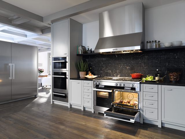 view of a modern kitchen with Miele appliances built into white cabinetry. Appliances are stainless steel and include a double oven range, a range hood, a double wall oven with warming drawer, and a large refrigerator.