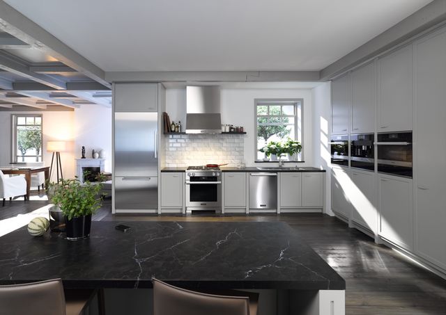 front view of a large kitchen with white walls and stainless steel appliances including a refrigerator, range, range hood, dishwasher, wall oven, and espresso machine