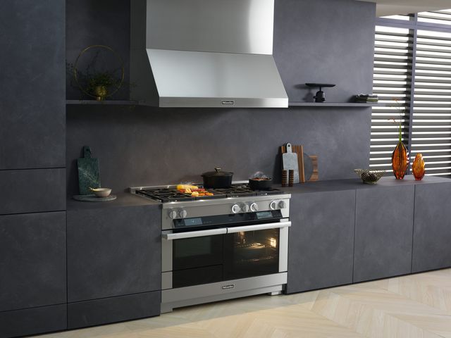 front view of a miele range in a contemporary dark kitchen with grey counters, walls, and backsplash. The oven light is on showing a pan inside, and there are various foods cooking on top