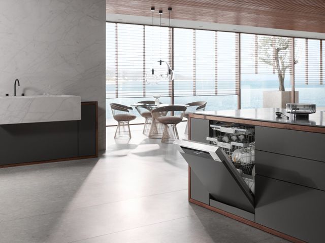 front view of a contemporary kitchen with an island on the left, featuring a partially opened built in dishwasher, and a sink against a marble wall on the left. The background shows an open-concept dining area with modern furniture and large windows for walls, with the ocean outside