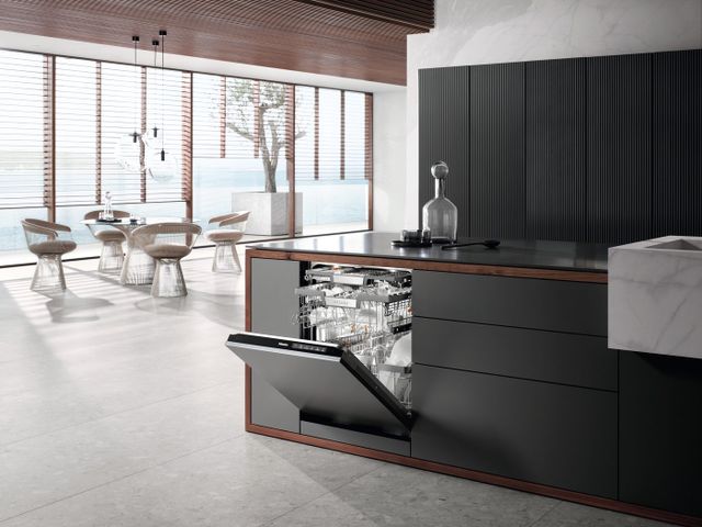 front view of an opened panel ready miele dishwasher, stocked with glassware and dishes. The dishwasher is built into a grey and black kitchen counter in a sleek, contemporary, home. On the left of the dishwasher in the background is an open concept dining room with large windows and a round table.