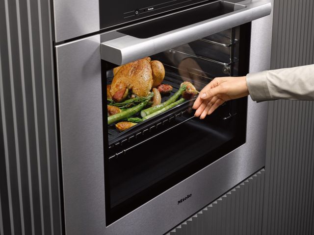 Close view of a hand pressing the glass of a Miele wall oven with a chicken baking inside