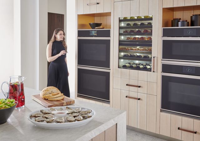 front view of a woman standing in a modern kitchen with two Monogram double ovens and a wine cooler between them built into beige panel cabinets. In the foreground is the corner of the kitchen island, with bread, oysters, and a pitcher of drink on top.