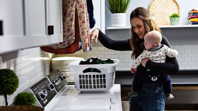 Front view of a modern laundry room and a woman holding a baby in front of a Speed Queen top load washer and dryer