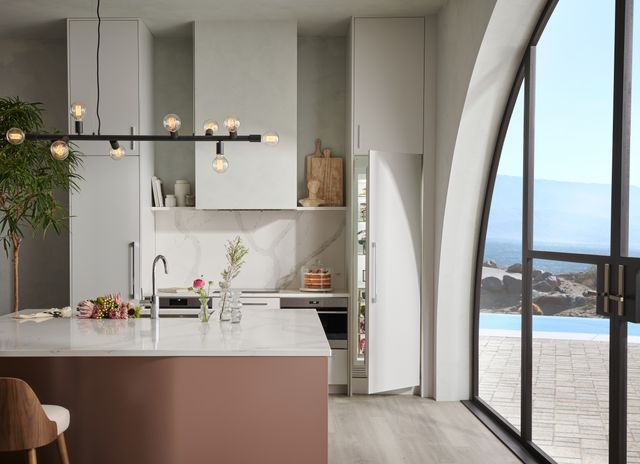 Front view of a modern kitchen with white walls and cabinetry, an island with a sink, two Wolf wall ovens built behind the island, and a Sub-Zero panel ready column refrigerator that is slightly opened on the right.