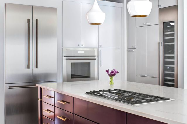 front view of a white kitchen with a stainless steel thermador french door refrigerator and matching appliances in the background and an island in the foreground