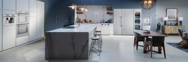 Wide view of a modern kitchen with stainless steel Thermador appliances, an island, and an open concept layout that leads into the living room