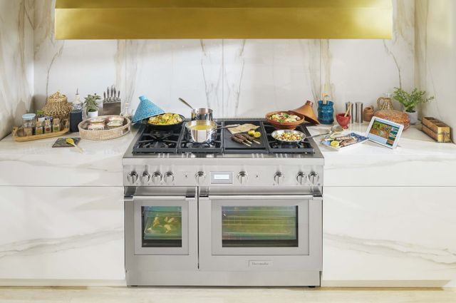 Front view of a modern kitchen with a marble backsplash and a Thermador range with breakfast cooking on it.