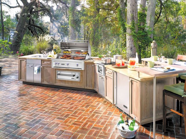 front view of a large modern outdoor kitchen with beige countertops and stainless steel appliances, including a large built in grill with various meats cooking on it