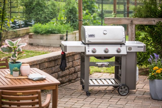 front view of a stainless steel weber freestanding grill on an outdoor patio with a garden in the background