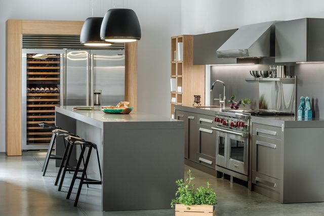 front view of a modern luxury kitchen with grey walls, light wood and grey cabinetry, and stainless steel Wolf and Sub-Zero appliances