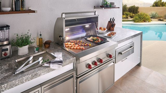 View of a kebabs cooking on a Wolf outdoor grill in a covered outdoor kitchen by a pool.
