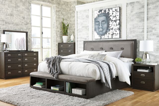 front view of a modern bedroom with white brick walls, light wood floors, and a dark brown modern bedroom furniture set with a storage bed, nightstand, dresser, and chest