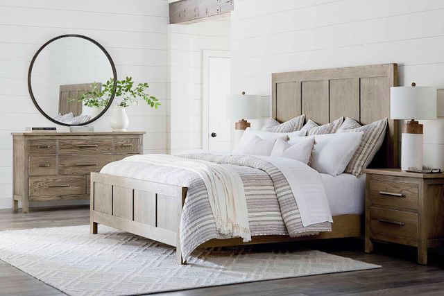 view of a modern bedroom with white panel walls and a light wooden modern bedroom set including a full bed, two nightstands (one out of view behind the bed, with a lamp peeking over), and a dresser