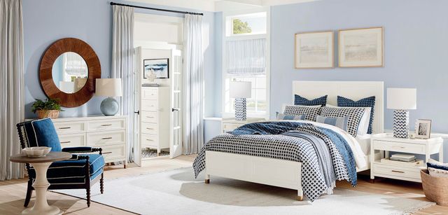 front view of a modern bedroom with light blue walls, light wooden floors, and a white modern bedroom set that includes a bed, two nightstands, a dresser, and a chest in the walk in closet in the background