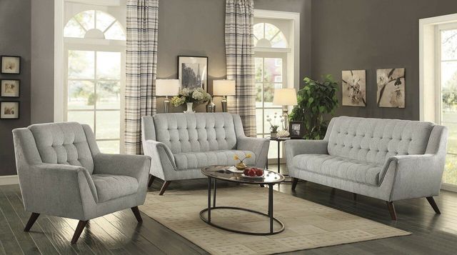 front view of a contemporary living room with Coaster furniture, including a light grey sofa, loveseat, and chair, a round glass coffee and side table, an area rug, and various home accents