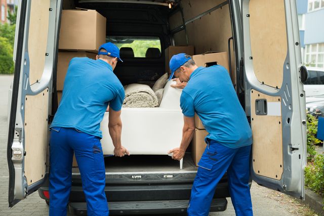 front view of two men wearing blue unloading a white sofa from the back of a furniture delivery truck