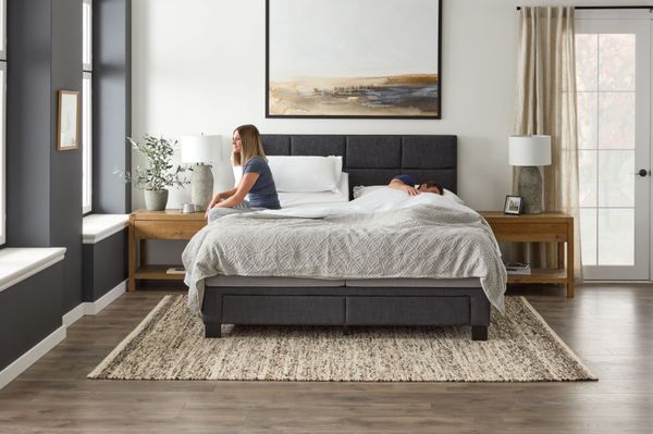front view of a woman sitting up on a malouf mattress and a man sleeping next to her, in a modern bedroom with a dark grey bed frame and white walls