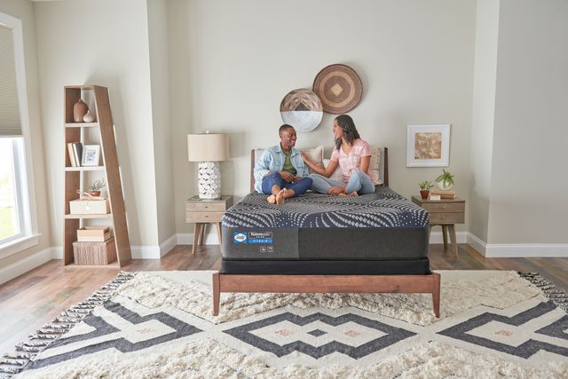 front view of a man and woman sitting and smiling on a sealy mattress in a boho bedroom