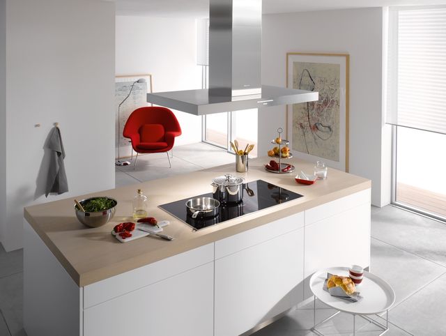 view of a large open kitchen and living room, with the kitchen in the foreground and the living room in the background. In the kitchen in a white island containing a black miele induction cooktop surrounded by cooking items