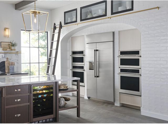 view of open white kitchen with gold accents. Wall has a large monogram refrigerator surrounded by monogram ovens on both sides. in view on the left is a kitchen island with a white marble countertop, and a built in wine cooler.